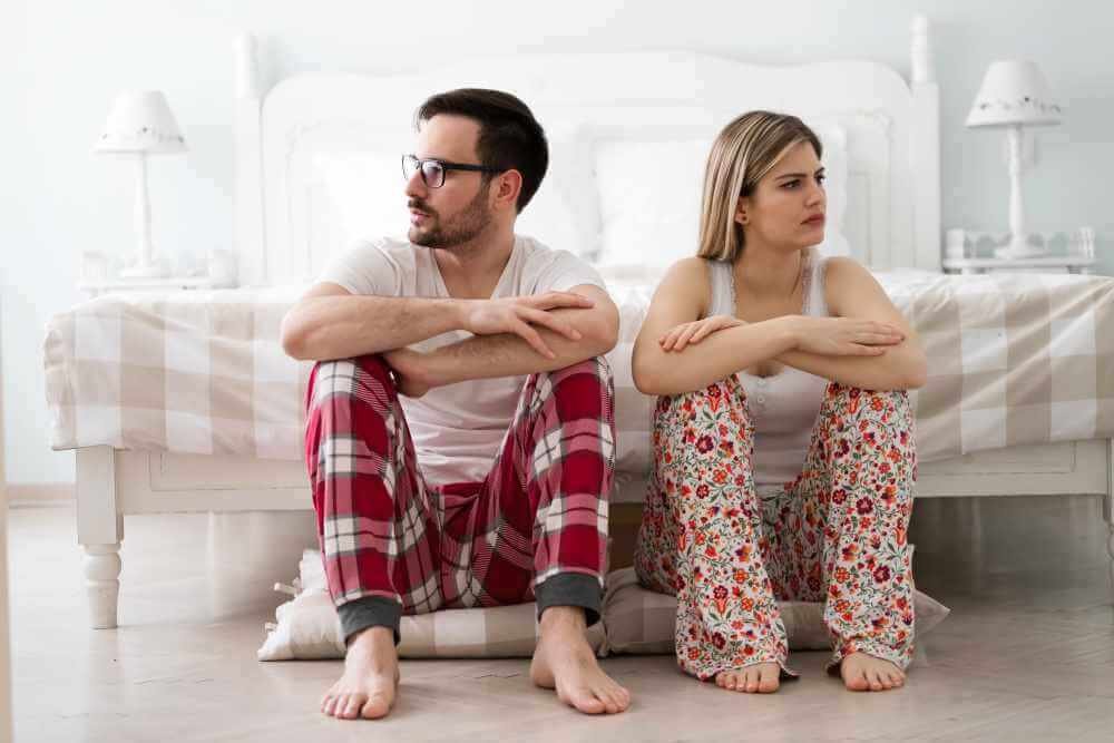 Man and woman in pajamas sitting at the end of their bed with their arms folded and looking away from one another